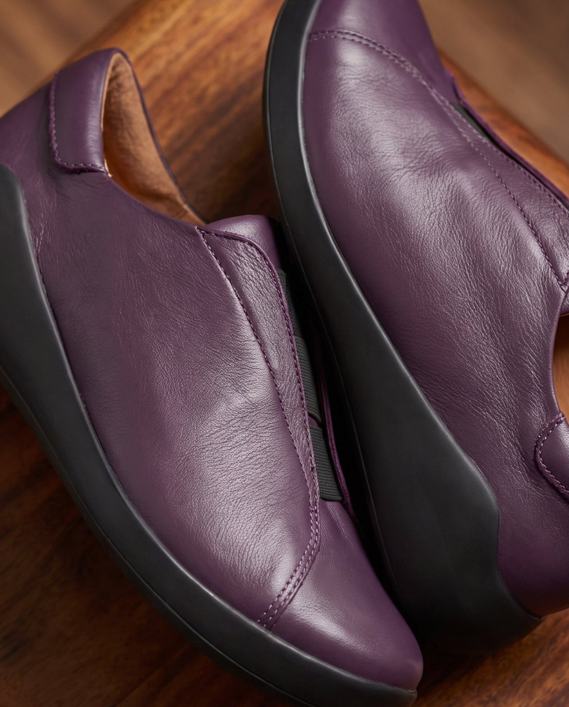 A pair of women's slip-on loafers in purple leather with black soles, displayed on a dark wooden surface.